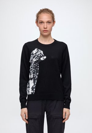 Le modèle porte un pull noir orné d’un grand motif de léopard blanc et un pantalon cargo noir, sur un fond clair uni.