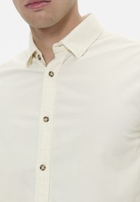 Camicia da uomo a bottoni in crema realizzata in tessuto testurizzato, con colletto a punta e multiple bottoni arrotondati marroni lungo la parte anteriore.