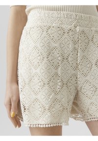 Krajkové šortky v krémové barvě s jemnými diamantovými vzory a zvlněným lemem s pom-pom detaily. Elastický pas pro pohodlí.