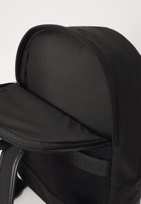 Zaino nero con un'esterno liscio e morbido. Presenta un'apertura con zip, una parte superiore arrotondata e inserti in rete sui lati per la ventilazione.