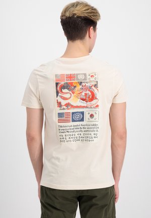 Osoba ubrana w beżowy t-shirt z czerwonym smokiem, flagami amerykańską, brytyjską, ONZ i południowokoreańską oraz tekstem w języku angielskim i koreańskim na plecach.
