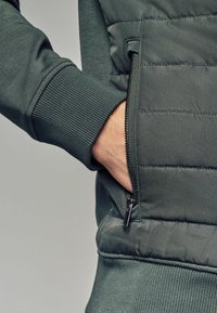 Dunkelgrüne gepolsterte Weste mit gerippten Bündchen, ausgestattet mit einer Reißverschlusstasche auf der linken Seite und einem strukturierten Stoff im gesteppten Muster.
