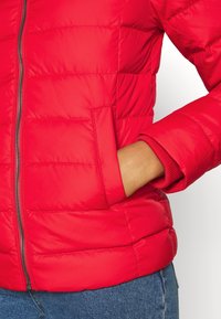 Main dans la poche d'une veste matelassée rouge avec une fermeture éclair visible à l'avant et un jean bleu.