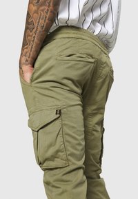 Cargo-Hosen in olivgrün, ausgestattet mit mehreren Taschen, einem elastischen Bund und einer glatten Textur. Detaillierte Nähte und ein Logo-Akzent sind vorhanden.