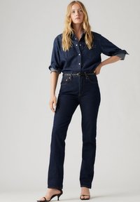 Donkerdenim shirt met opgerolde mouwen, twee borstzakken; gecombineerd met hoge donkere spijkerbroeken en zwarte haksandalen. Eenvoudig, klassiek ontwerp.