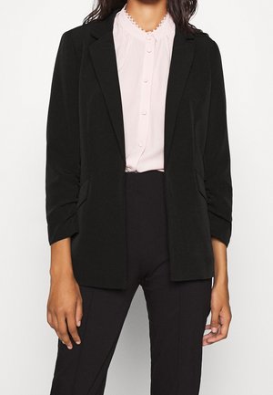 Femme portant un blazer noir cintré sur un chemisier rose clair boutonné et un pantalon noir ajusté sur fond blanc.