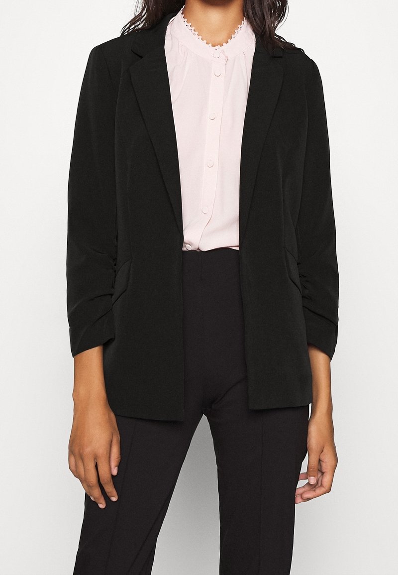 Femme portant un blazer noir cintré sur un chemisier rose clair boutonné et un pantalon noir ajusté sur fond blanc.