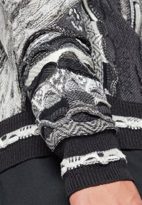 Schwarzer und grauer gestrickter Pullover mit strukturierten Mustern, geschichteten Nähten und kontrastierenden weißen Akzenten an den Bündchen und dem Saum.