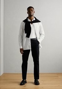 Cămașă albă cu dungi, purtată peste un tricou alb, pantaloni negri și mocasini negri. O puloveră neagră este aruncată pe umeri.