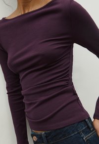 Maglietta a maniche lunghe in un profondo viola, con scollo a barca rilassato e dettaglio arricciato sui lati. Texture liscia con una silhouette aderente.