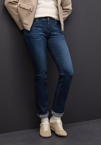 Persona con chaqueta beige, suéter blanco, jeans azul arremangados, calcetines blancos y zapatillas beige de pie contra una pared gris oscuro.