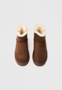 Bottines en suède marron avec une douce doublure en fausse fourrure crème, bout rond et semelle en caoutchouc texturé. Présente un bord festonné en haut.