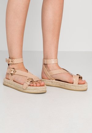 Sandalias espadrilles de color rosa claro con textura tejida, correas en el tobillo y herrajes metálicos. Cuentan con una suela texturizada y diseño de punta abierta.