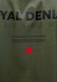 Alyvuogių žalio medvilninio marškinėlio dizainas su ryškiai juodu tekstu "ROYAL DENIM DIVISION" ir raudonu apskritimo akcentu apačioje. Minkšta tekstūra, apvalus kaklas.