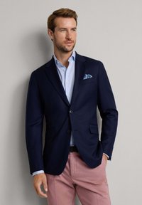 Blazer navy con due bottoni, tasche laterali, abbinato a una camicia azzurro chiaro e pantaloni rosa chiaro, con un fazzoletto da taschino a fantasia.