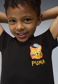 Mali dječak s osmijehom, ruku iza glave, u crnoj majici s printom žutog mačka s plavom kapom i tekstom "PUMA".