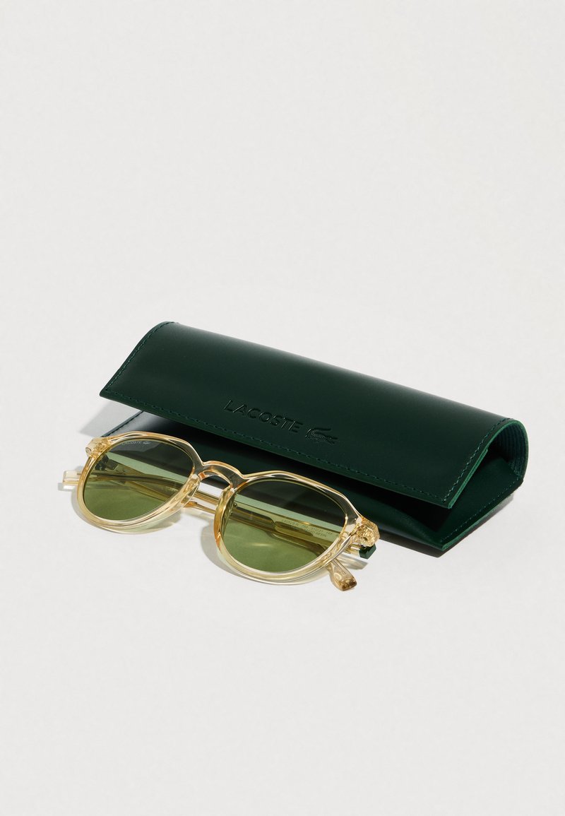 Lunettes de soleil à monture jaune transparente avec des verres verts posées à côté d'un étui à lunettes Lacoste vert foncé fermé sur une surface blanche.