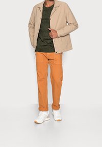 Veste beige, pull en tricot vert foncé, pantalon orange avec poches, et baskets blanches avec des accents orange. Tissu lisse et coupe structurée.