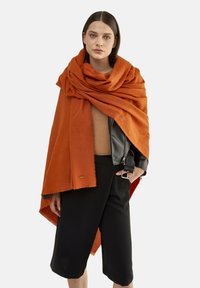 Orange uldet wrap med en blød tekstur, der har frynser i kanterne. Båret over en brun top og en sort jakke, kombineret med sorte shorts.