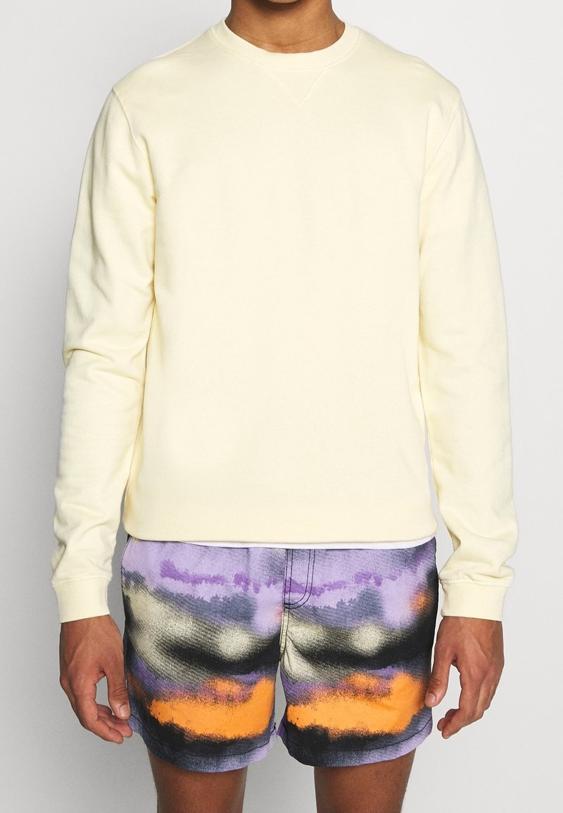 Sweatshirt giallo chiaro con scollo a girocollo, abbinato a pantaloni corti a motivi viola e neri con un accento arancione. Tessuto morbido e vestibilità casual.