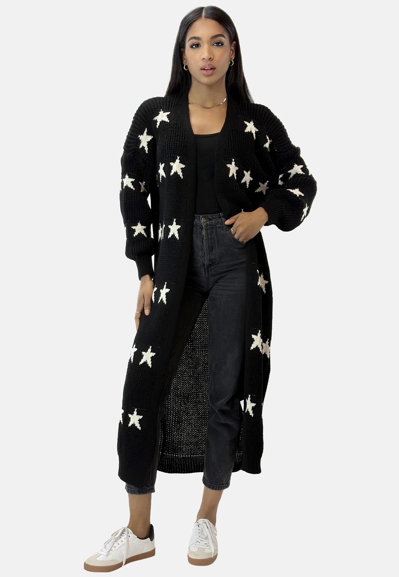 Gilet en tricot noir avec un motif d'étoiles blanches, manches longues, devant ouvert et longueur genoux, associé à un jean noir et des baskets.