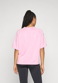 Kvinne med langt flettehår iført en løs rosa t-skjorte med korte ermer og svarte leggings, vendt bort fra kameraet mot en enkel lys bakgrunn.