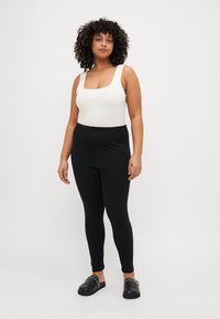 Zwarte legging gemaakt van rekbaar materiaal, gecombineerd met een witte tanktop met een vierkante halslijn. Het model staat tegen een effen achtergrond.