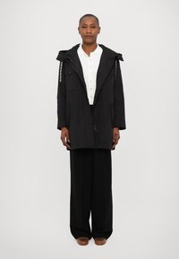Parka noir imperméable avec capuche, fermeture éclair, poches latérales et cordon de serrage ajustable. Texture lisse, associé à une chemise blanche boutonnée et un pantalon noir.