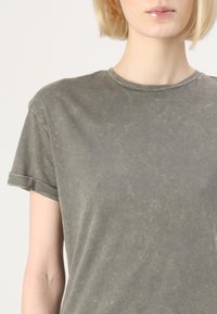 Oliwkowa bawełniana koszulka z okrągłym dekoltem, krótkimi rękawami i podwiniętymi mankietami. Materiał ma subtelną, wyblakłą fakturę i wzór.