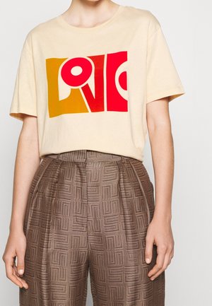 T-shirt beige avec un graphisme abstrait "LOVE" rouge et jaune moutarde, associé à un pantalon marron texturé à motifs taille haute.