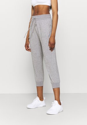 Szczupła osoba ubrana w szare spodnie dresowe ze sznurkiem i białe trampki, stojąca na jednolitym białym tle.