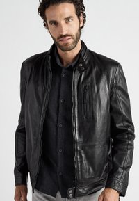 Schwarze Lederjacke mit Reißverschluss, Stehkragen, Brusttasche und Seitentaschen. Strukturierte Oberfläche mit eleganten Nähten.