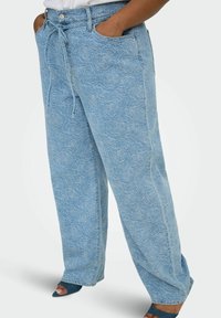 Jean large bleu clair avec motif de rose en relief, taille à cordon, poches avant, porté avec des talons ouverts teal foncé.