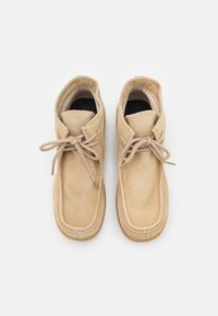 Mocassins en daim beige avec des bouts ronds et des lacets beige noués en nœuds, vus de dessus sur un fond blanc.