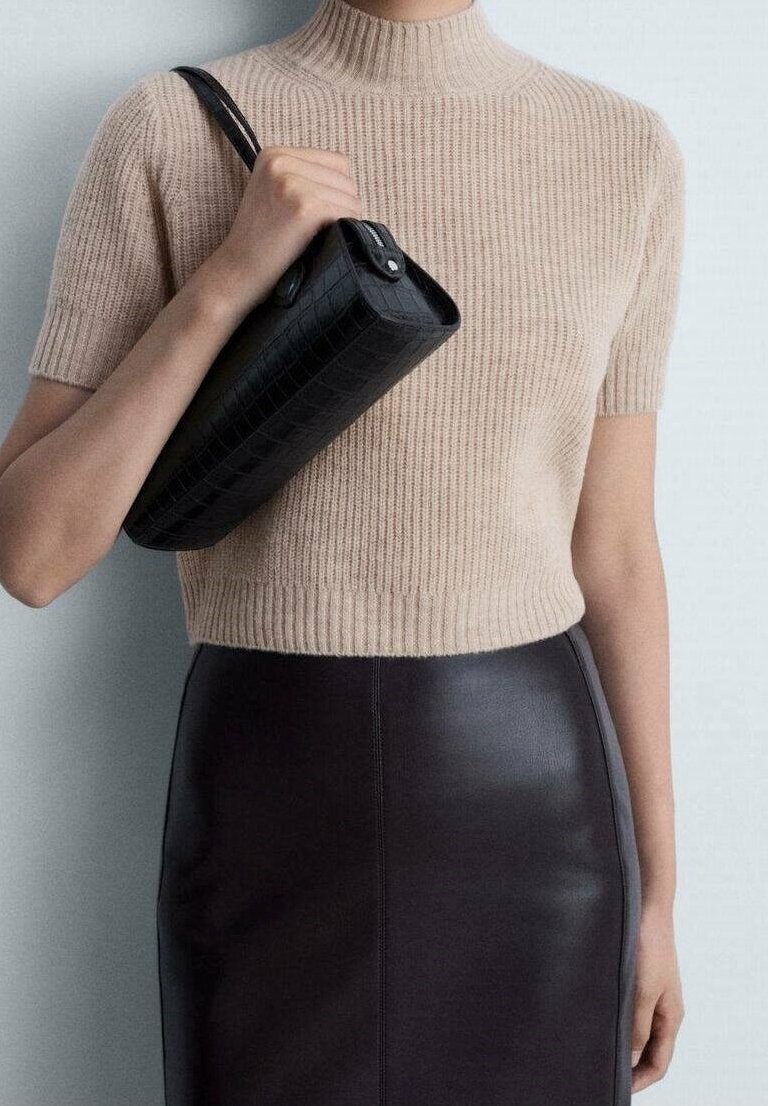 Personne portant un haut en maille beige à manches courtes et une jupe en cuir noire, tenant un sac à main noir texturé sur une épaule.