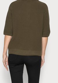 Olivově zelený pletený svetr s krátkým rukávem, ribbovanou strukturou a uvolněným střihem, kombinovaný s černými slim džínami.