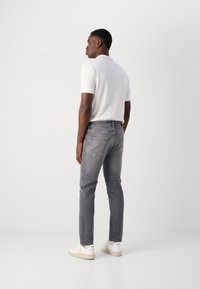 Jeans gri cu o croială dreaptă, fabricat dintr-un amestec de bumbac, cu cinci buzunare și un tricou polo alb. Modelul poartă adidași albi, văzut din spate.