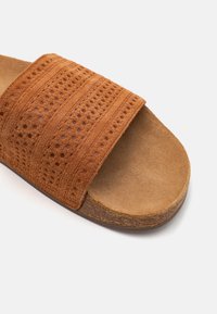 Sandalo scivolato in suede marrone con una fascia ampia e testurizzata caratterizzata da perforazioni, e una pianta piatta color beige per il comfort. Punta arrotondata e tacco basso.
