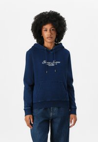 Sweat à capuche bleu marine avec une poche avant, un cordon de serrage et le texte blanc "Guess Jeans American Tradition California" sur la poitrine.