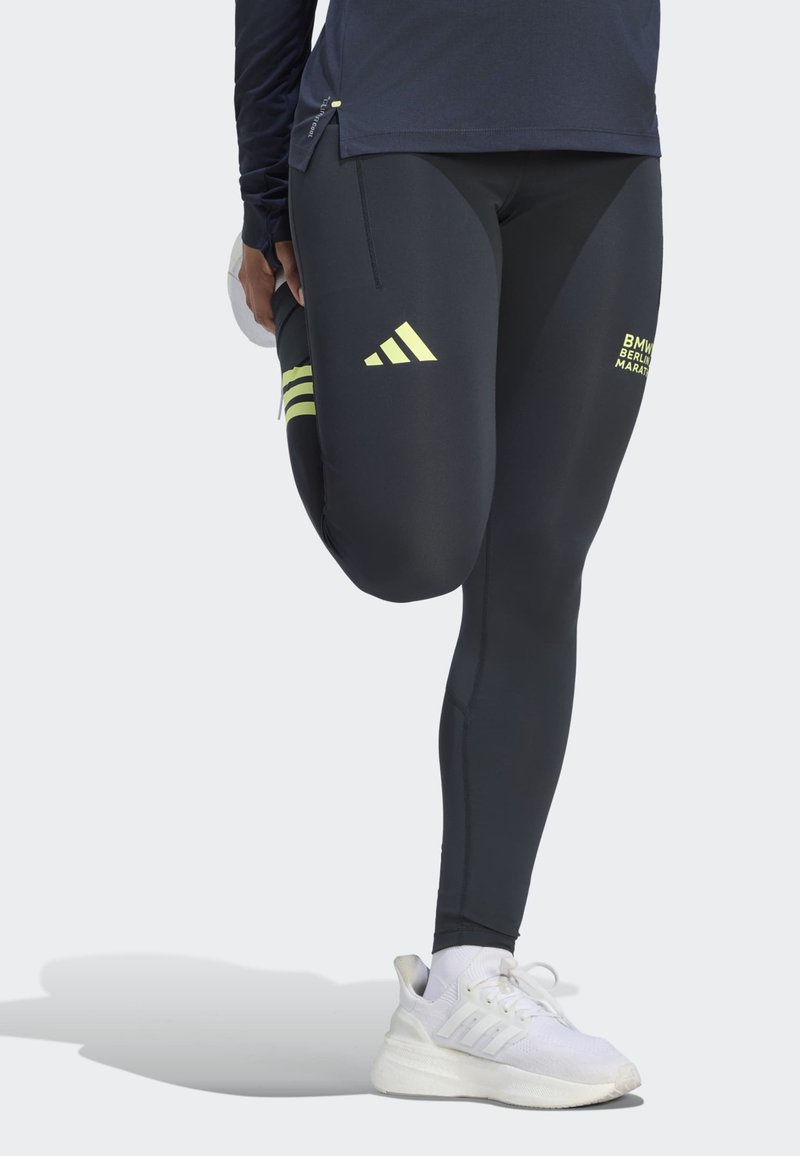 Човек, облечен в черни клинове Adidas с логото на Берлинския маратон, разтяга крака си, докато носи бели маратонки.
