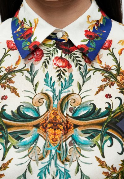 Košeľa s kvetinovým vzorom so bielym základom, živými červenými a zelenými kvetmi, modrým golierom a zložitými ozdobnými detailmi pozdĺž prednej časti.