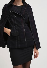 Cape veste à carreaux noirs avec col, fermeture éclair sur le devant et poches latérales. Le tissu a une texture douce et présente des accents rouge foncé.