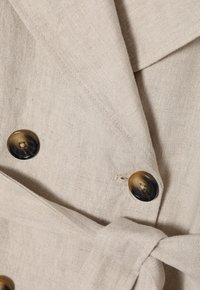 Beige linen coat with a wrap design, featuring two large, dark brown buttons and a fabric belt. Textured surface with visible weave.