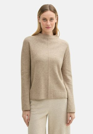 Pull en tricot beige avec un col montant, des épaules tombantes et une texture subtile. Associé à un pantalon beige clair pour un look coordonné.