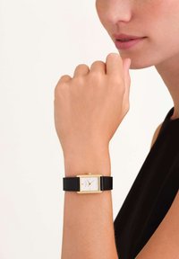 Orologio d'oro rettangolare con quadrante bianco e cinturino in pelle nera, indossato su una mano. Presenta un design minimalista e indicatori delle ore semplici.