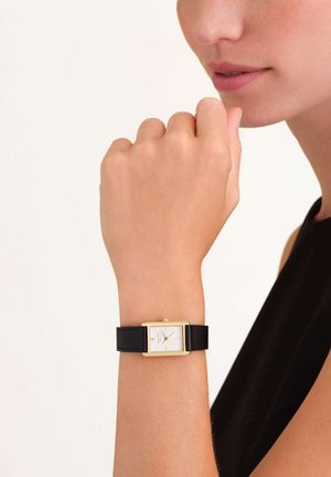 Montre en or rectangulaire avec un cadran blanc et un bracelet en cuir noir, portée sur une main. Présente un design minimaliste et des index horaires simples.