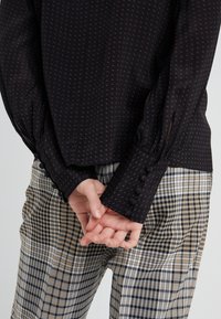 Svart blus med ett texturerat mönster, långa ärmar med knappdetaljer, matchad med rutiga byxor i beige, svart och vitt.