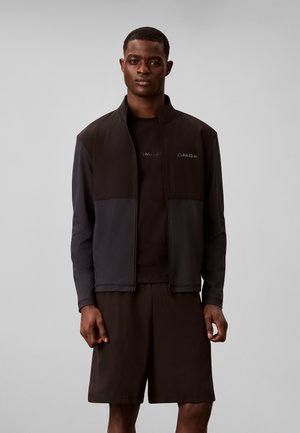 Homme portant une veste zippée noire par-dessus un t-shirt noir et un short noir, debout devant un fond gris.