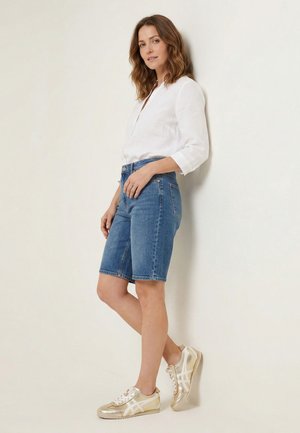 Frau lehnt an einer weißen Wand, trägt ein weißes Langarmshirt, blaue Jeans-Shorts bis zum Knie und gold-weiße Turnschuhe.