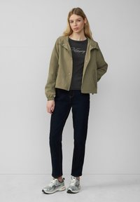 Olijfgroen lichtgewicht jack met een capuchon, knopen aan de voorkant en elastische manchetten, gedragen over een donkere t-shirt en gecombineerd met donkere jeans en zilveren sneakers.
